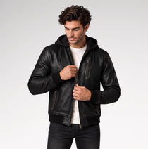 A man wearing a black leather hooded jacket with zippered pockets, styled over a white t-shirt and dark jeans, photographed against a light gray studio background.