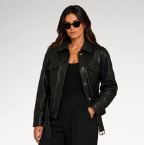 A woman modeling a black leather jacket with a matching black belt, styled with a black top and trousers against a light gray studio background.