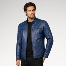 A man wearing a blue leather jacket over a black shirt and black pants, standing against a light gray studio background.
