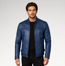 A man wearing a blue leather motorcycle jacket over a black t-shirt and black pants, standing against a light gray studio background.