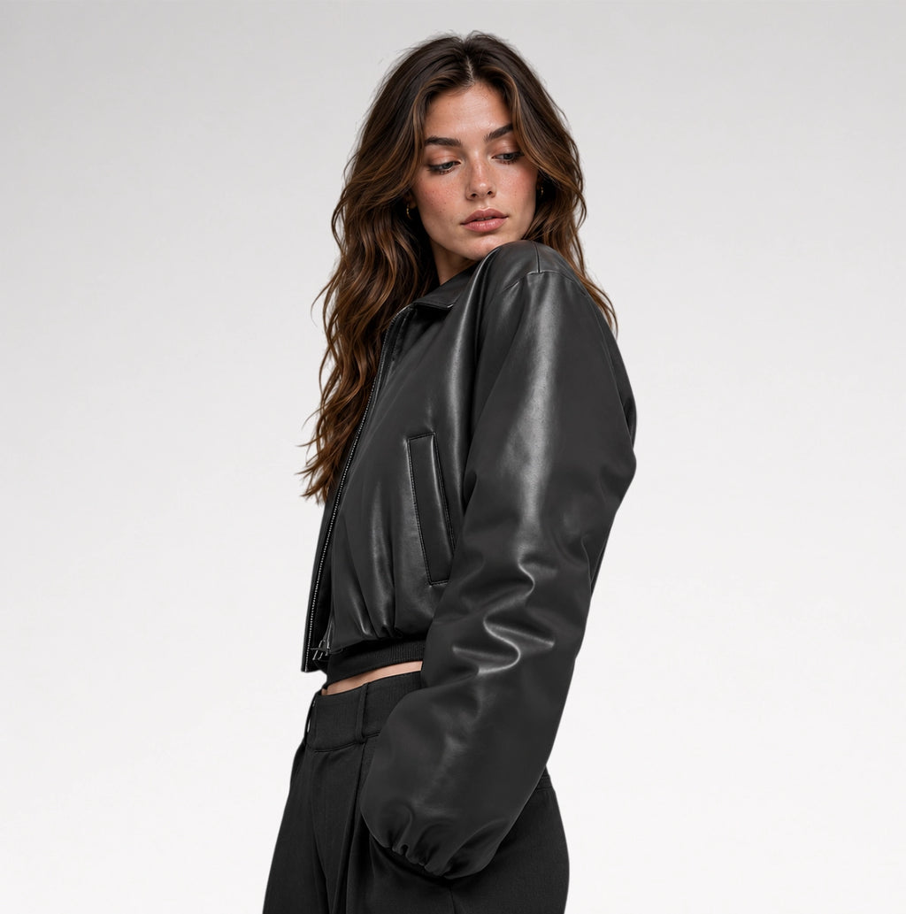 A woman wearing a black faux-leather cropped jacket with a collar and front zipper, paired with black trousers, photographed against a light gray studio background.
