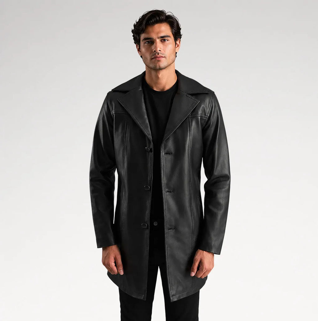 A man modeling a black leather trench coat over a black shirt and trousers, standing against a light gray studio background.