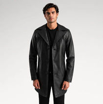 A man modeling a black leather trench coat over a black shirt and trousers, standing against a light gray studio background.