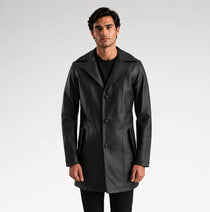 A man modeling a black leather-like coat over a black outfit, standing against a light gray studio background.