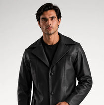 A man modeling a black leather-like coat over a black outfit, standing against a light gray studio background.