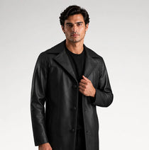 A man modeling a black leather-like coat over a black outfit, standing against a light gray studio background.