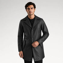 A man modeling a black leather-like coat over a black outfit, standing against a light gray studio background.