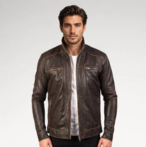 A man wearing a dark brown leather jacket with contrast stitching over a white t-shirt, photographed against a light gray studio background.