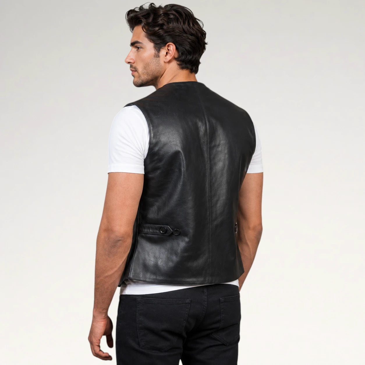 A man shown from the back wearing a black leather vest over a white t-shirt and black jeans, photographed against a light gray studio background.