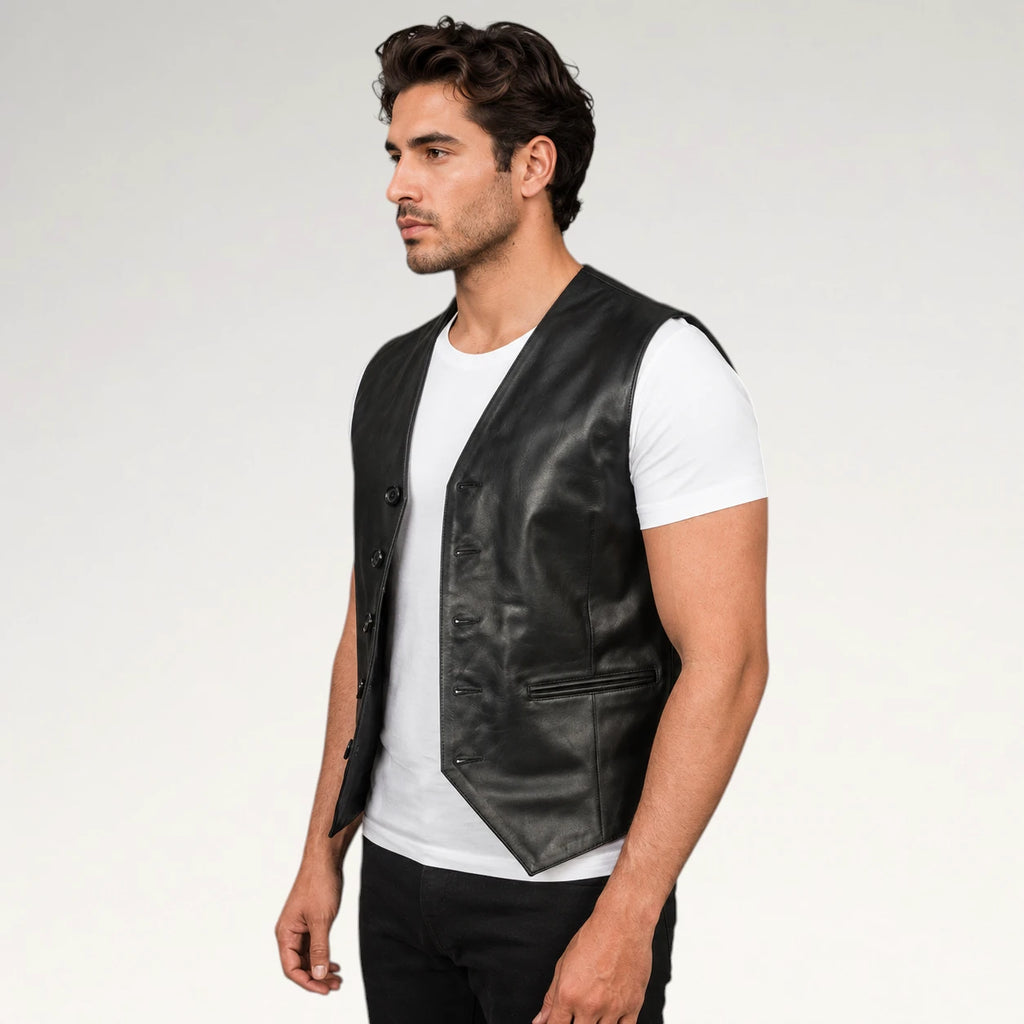 A man wearing a black leather vest over a white t-shirt and black pants, photographed from the waist up against a light gray studio background.