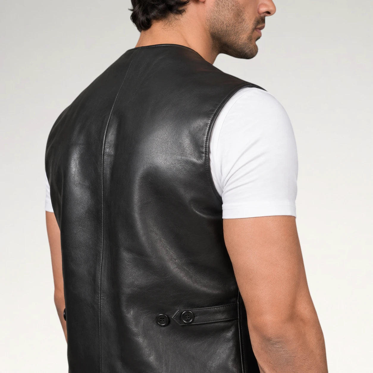 A man wearing a black leather vest over a white t-shirt, shown from a side-back angle against a light gray studio background.