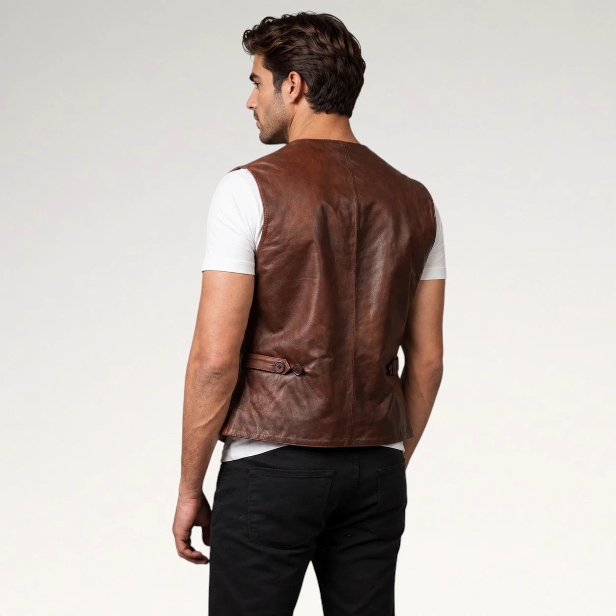 A man shown from the back wearing a brown leather vest over a white t-shirt and black pants, photographed against a light gray studio background.