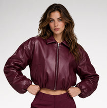A woman wearing a glossy, deep burgundy faux-leather cropped jacket and matching trousers, photographed from the side against a clean white studio background.
