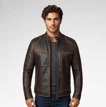 A man wearing a dark brown leather motorcycle jacket with contrast tan stitching, a dark gray t-shirt, and blue jeans, standing against a light gray studio background.