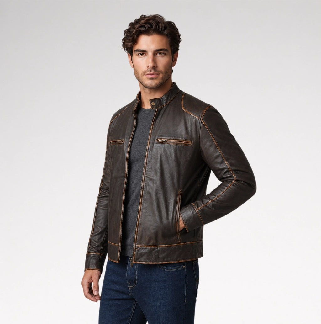 A man wearing a dark brown leather jacket with contrasting tan stitching, a dark gray t-shirt, and blue jeans, standing against a light gray studio background.