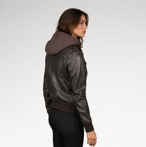 A woman shown from the side profile wearing a dark brown faux-leather jacket with an attached brown hood, paired with black pants, against a light gray studio background.