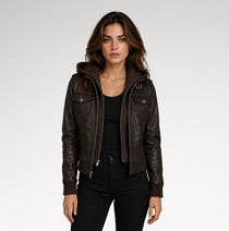 A woman modeling a dark brown faux-leather hooded jacket with a front zipper and flap chest pockets, paired with a black top and black pants, against a light gray studio background.