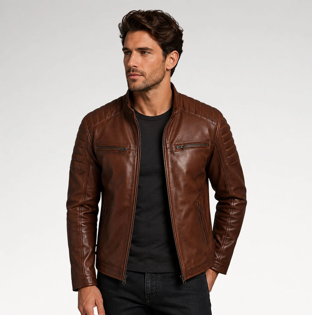 A man wearing a brown leather moto jacket over a black t-shirt and dark jeans, photographed against a light gray studio background.