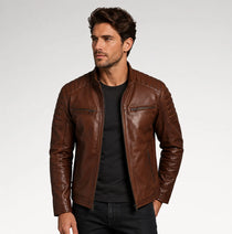 A man wearing a brown leather moto jacket over a black t-shirt and dark jeans, photographed against a light gray studio background.