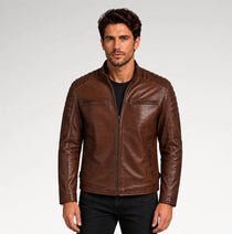 A man wearing a brown leather jacket with a front zipper, zippered chest pockets, and quilted shoulder detailing, paired with a black shirt and black jeans, against a light gray studio background.