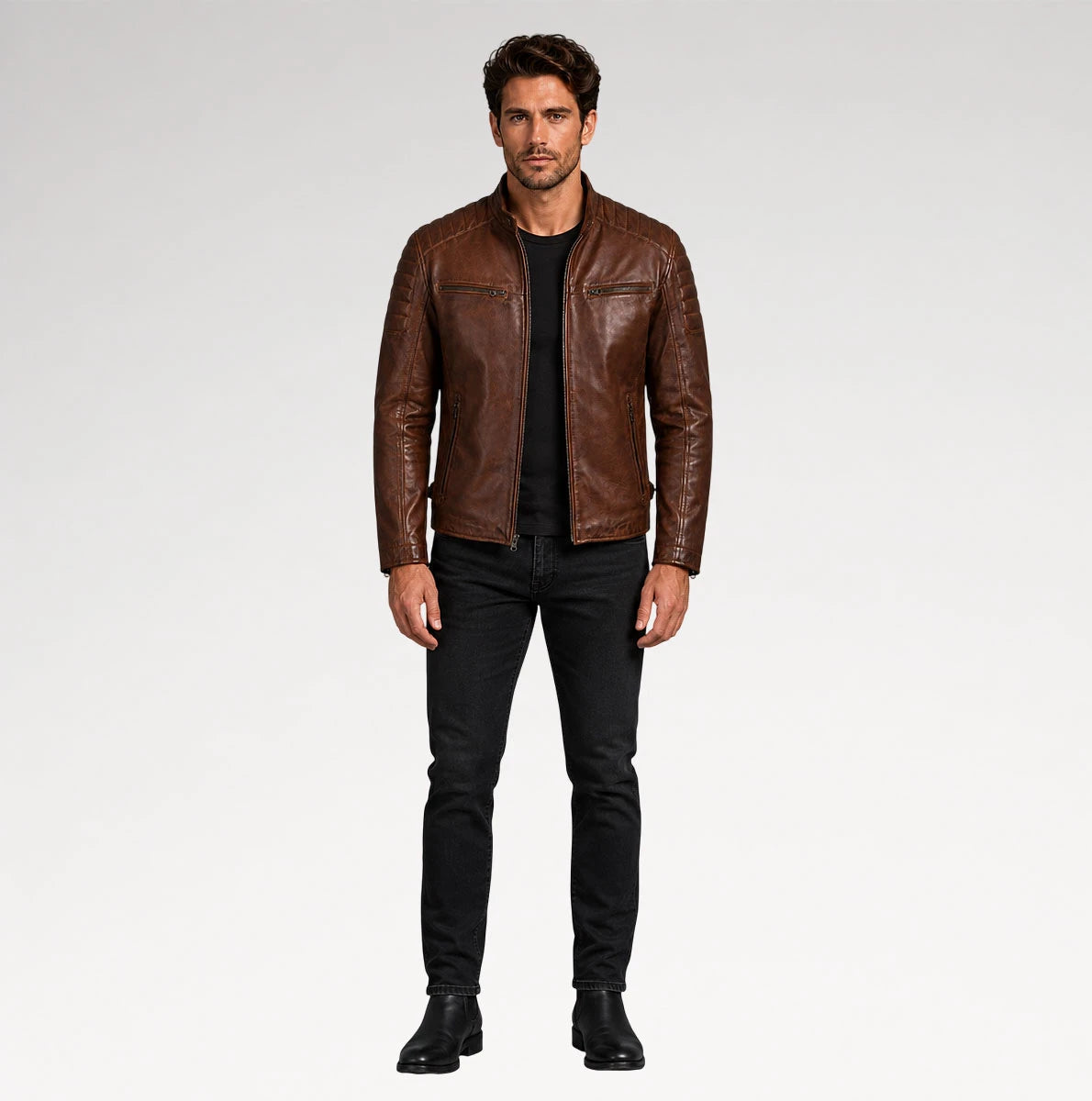 A man modeling a brown leather jacket, black t-shirt, black jeans, and black boots against a light gray studio background.