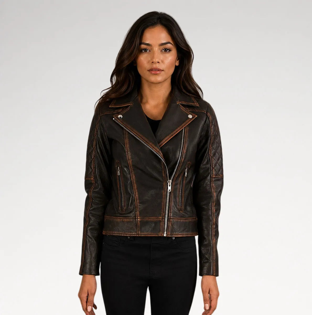 A woman modeling a dark brown distressed leather moto jacket with silver zippers and contrast stitching, paired with black pants against a light gray studio background.