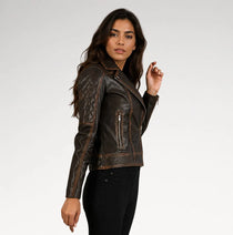 A woman modeling a dark brown distressed leather motorcycle jacket with quilted shoulder panels and zippered pockets, paired with black pants against a light gray studio background.
