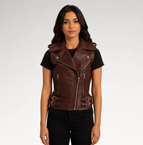 A woman modeling a dark brown faux-leather moto vest with silver hardware, worn over a black t-shirt and black jeans, against a light gray studio background.
