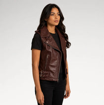 A woman modeling a dark brown faux leather moto vest with silver zippers and decorative side buckle details, worn over a black t-shirt and black pants against a light gray studio background.