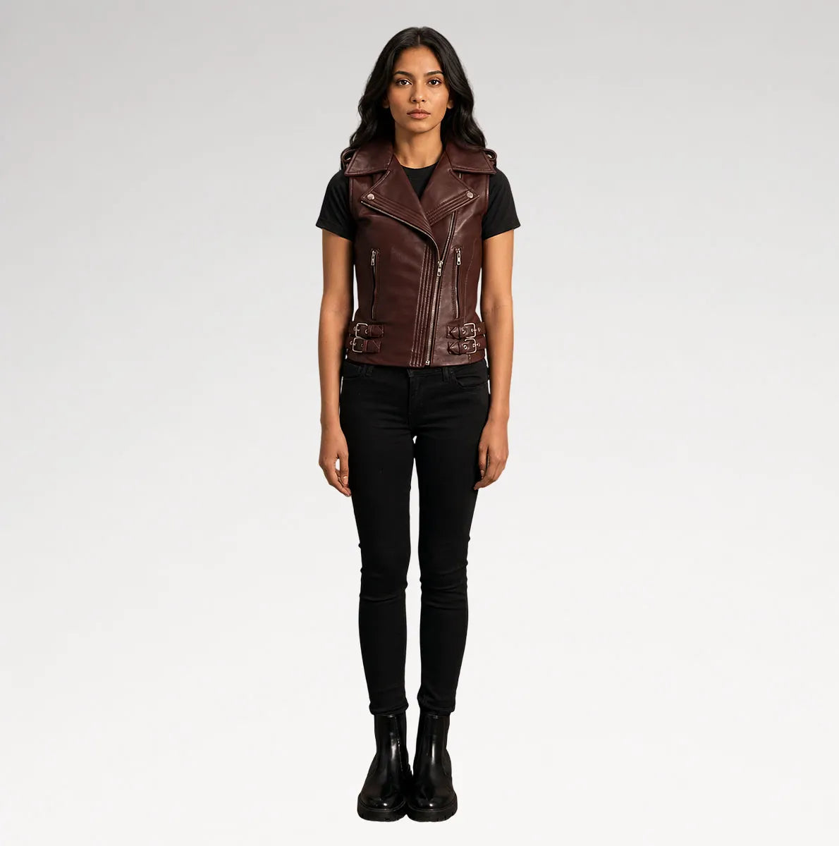 A woman modeling a dark brown faux-leather moto vest over a black t-shirt and black skinny jeans, standing against a light gray studio background.