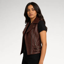 A woman wearing a dark brown faux leather moto vest over a black t-shirt and black jeans, photographed against a light gray studio background.