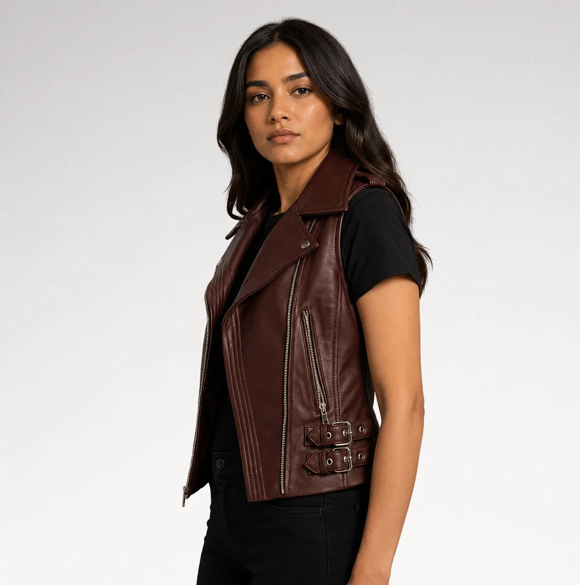 A woman wearing a dark brown faux leather moto vest over a black t-shirt and black jeans, photographed against a light gray studio background.