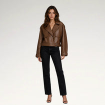A woman modeling a cropped brown leather moto jacket with black straight-leg jeans and black heeled sandals, standing against a plain light gray studio background.