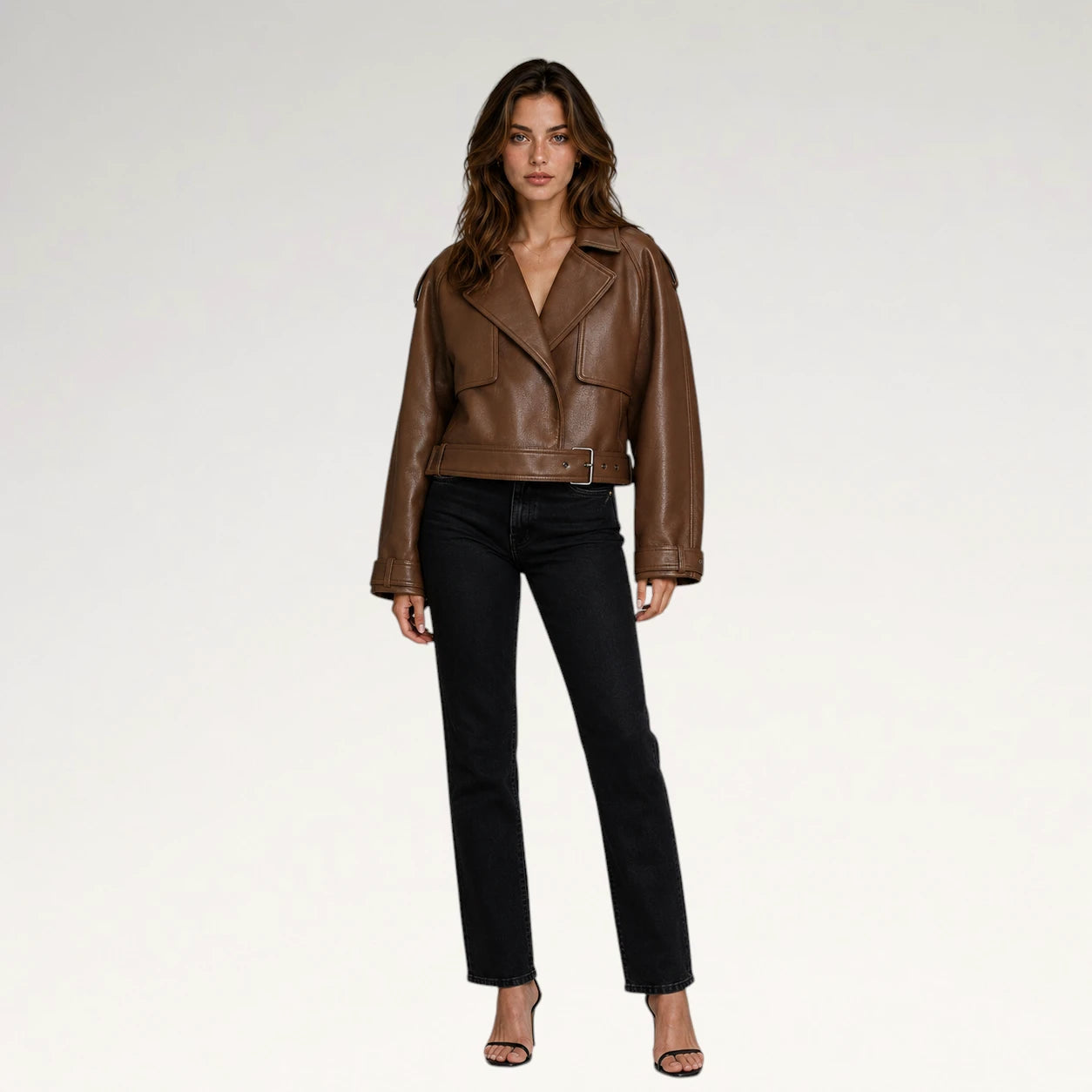 A woman modeling a cropped brown leather moto jacket with black straight-leg jeans and black heeled sandals, standing against a plain light gray studio background.