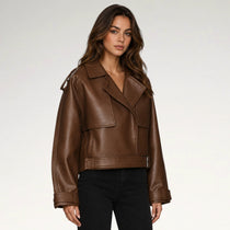 A woman modeling a cropped brown faux-leather moto jacket with a belted waist and asymmetrical collar, paired with black pants, against a light gray studio background.