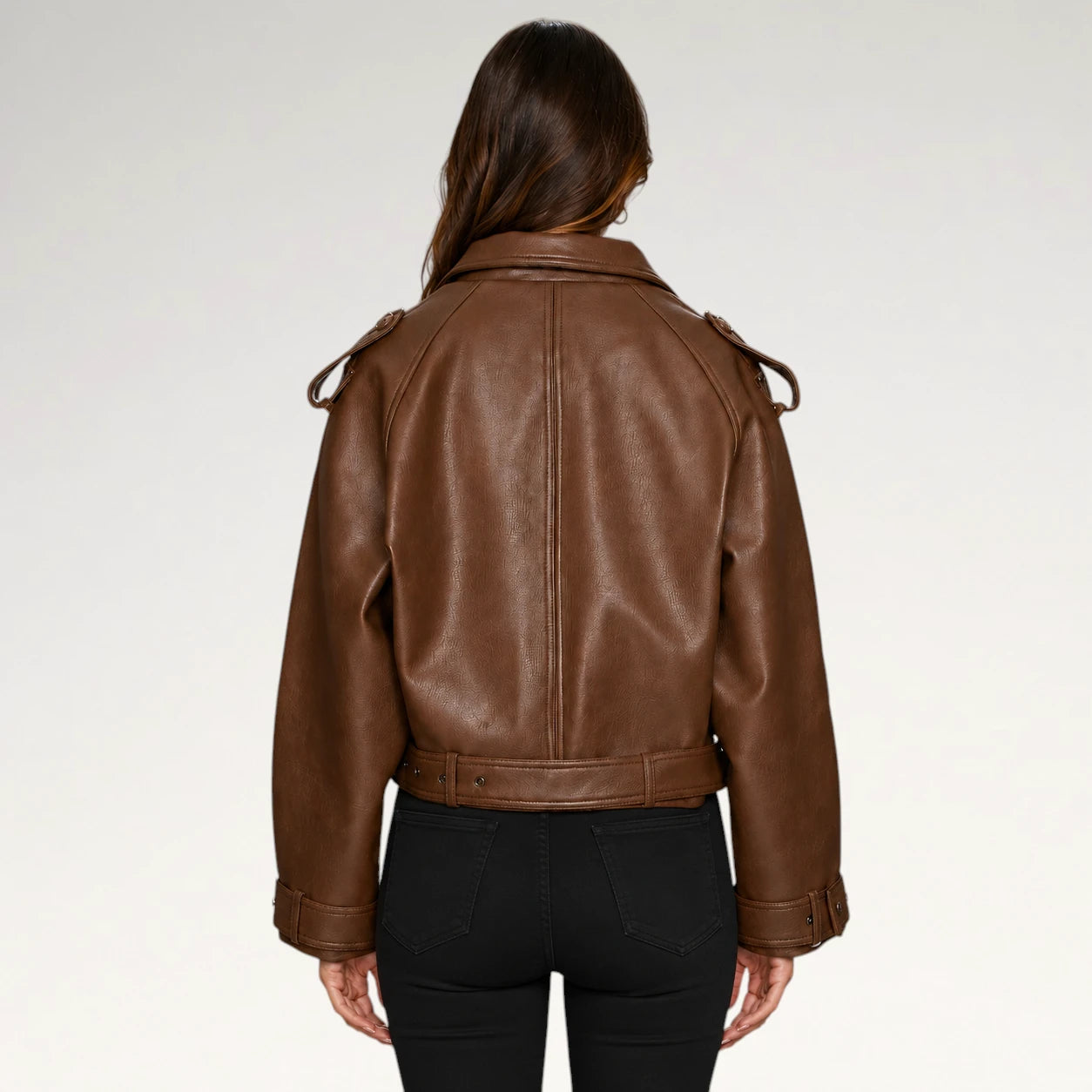 A woman viewed from the back wearing a brown leather jacket with shoulder epaulets and a matching belt, paired with black pants, against a light gray studio background.