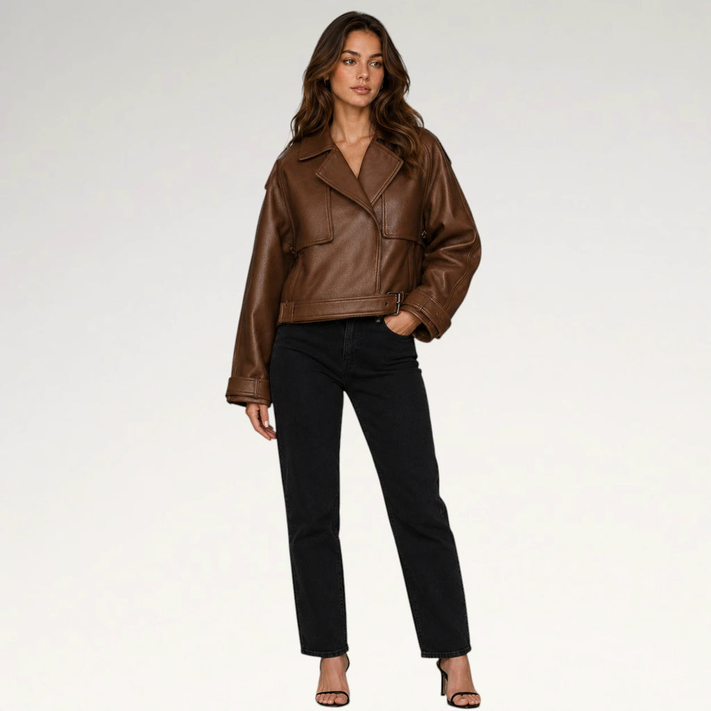 A woman modeling a brown faux-leather moto jacket with black pants and black sandals, standing against a light gray studio background.
