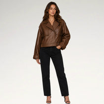 A woman modeling a brown faux-leather moto jacket with black pants and black sandals, standing against a light gray studio background.