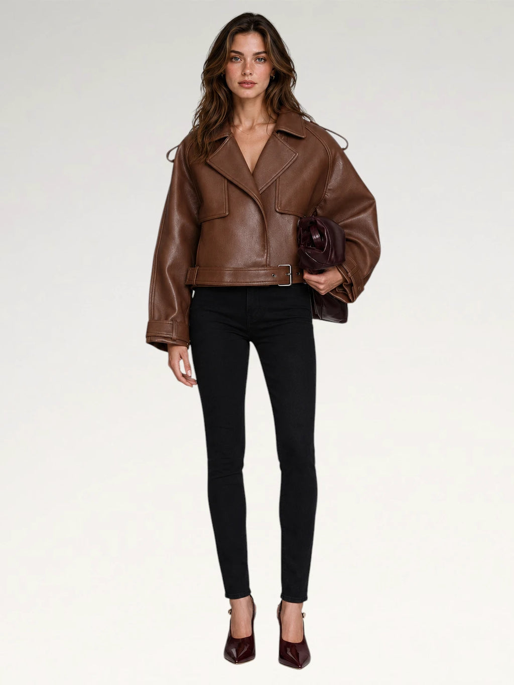 A woman modeling a brown leather moto jacket with a belted waist, paired with black skinny pants and burgundy heels, holding a dark brown clutch against a plain white studio background.