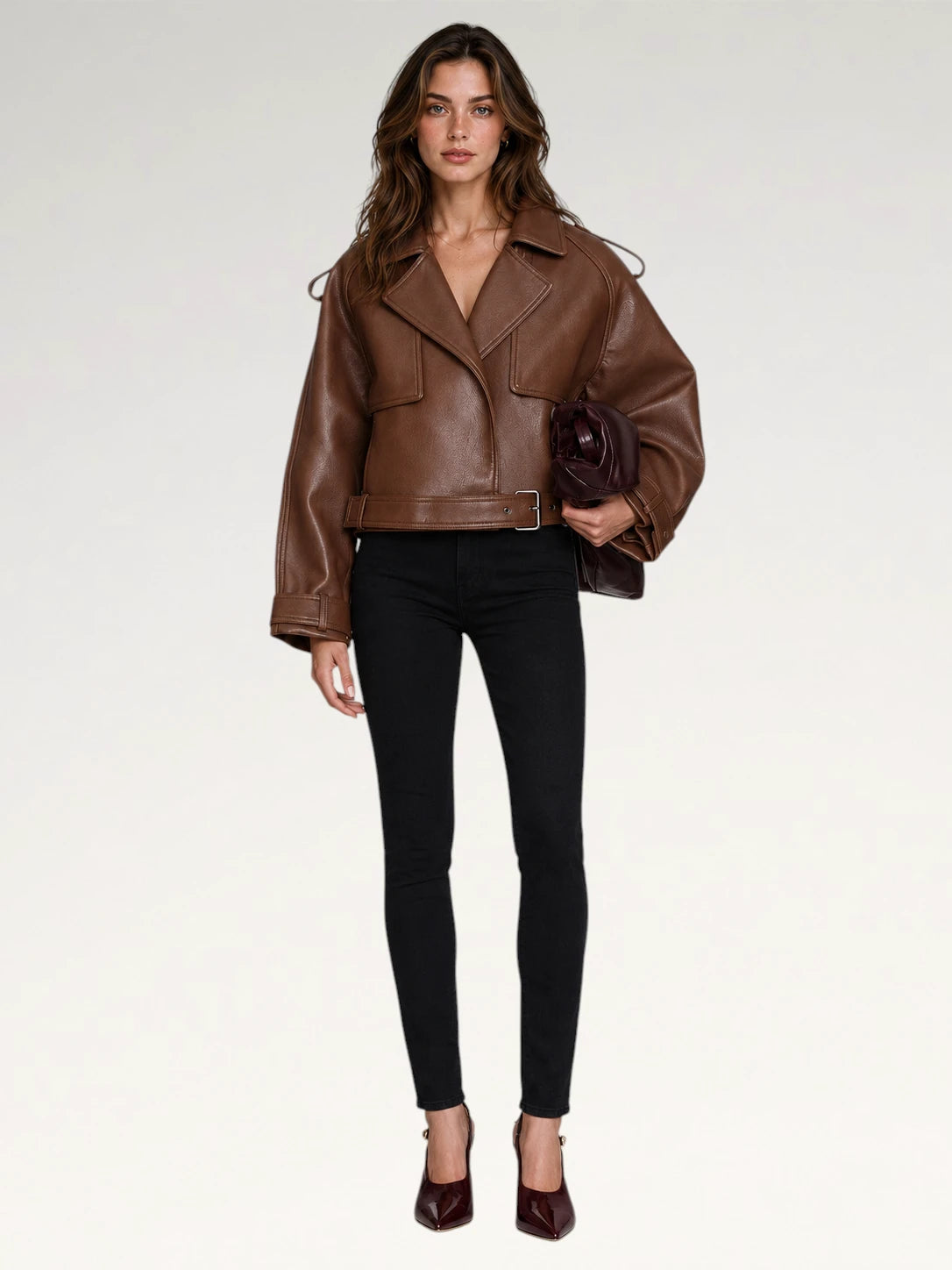 A woman modeling a brown leather moto jacket with a belted waist, paired with black skinny pants and burgundy heels, holding a dark brown clutch against a plain white studio background.