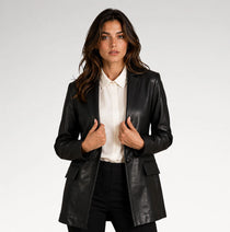 A woman modeling a black faux-leather blazer over a white collared shirt and black trousers, photographed against a light gray studio background.
