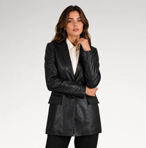 A woman modeling a black faux-leather blazer with a structured silhouette, worn over a white collared shirt, against a plain light gray studio background.