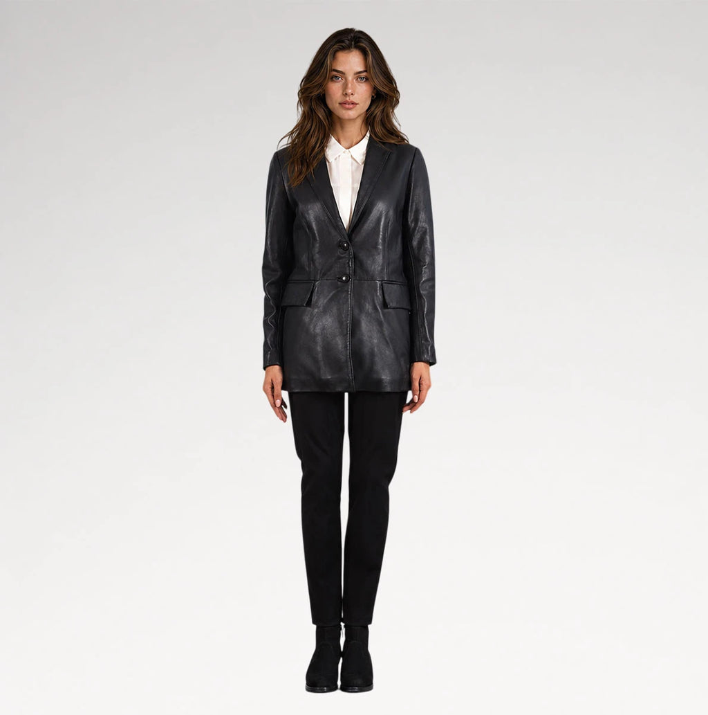 A woman modeling a black leather blazer over a white collared shirt, paired with black trousers and black ankle boots, against a light gray studio background.