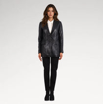 A woman modeling a black leather blazer over a white collared shirt, paired with black trousers and black ankle boots, against a light gray studio background.