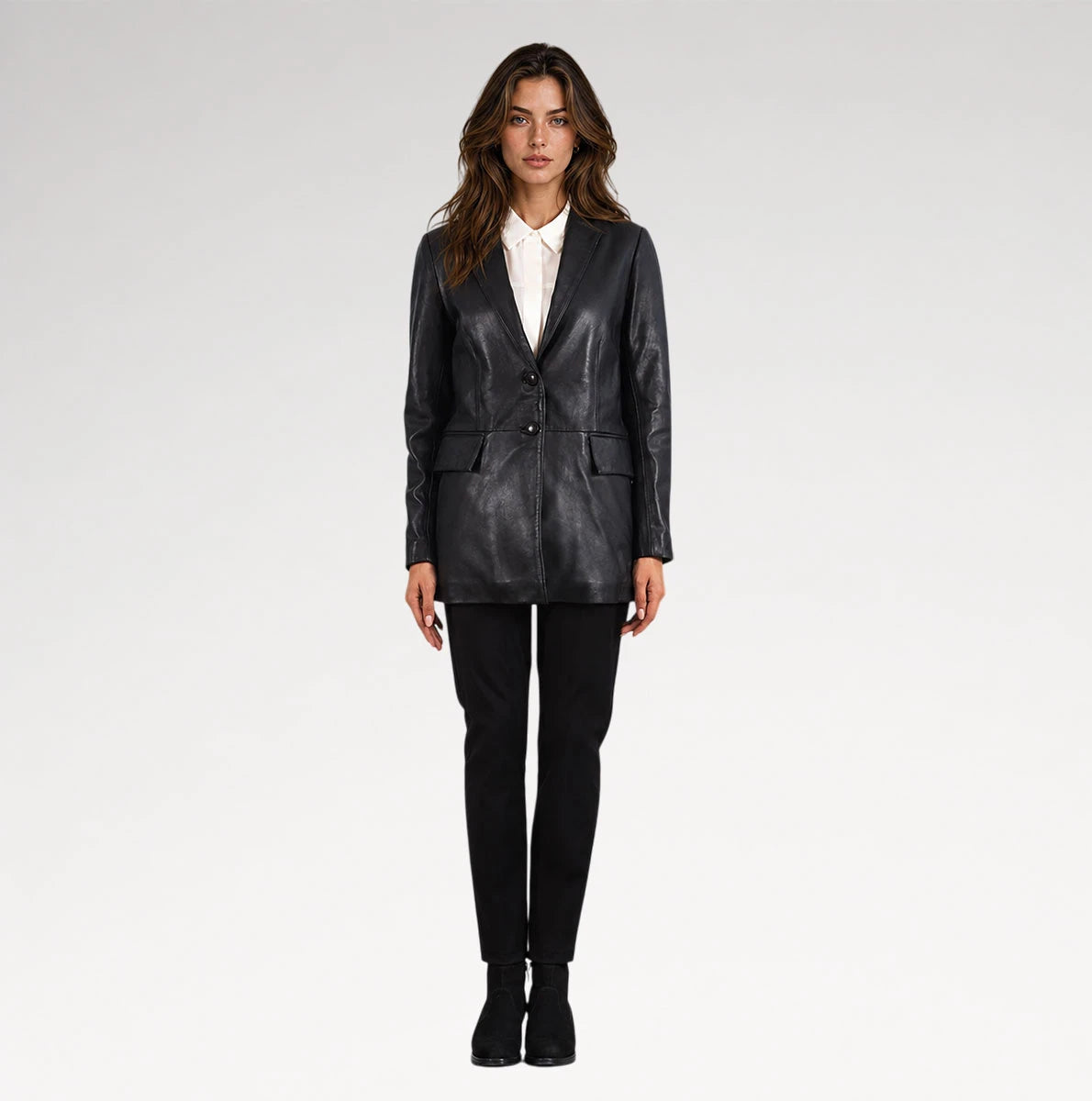 A woman modeling a black leather blazer over a white collared shirt, paired with black trousers and black ankle boots, against a light gray studio background.