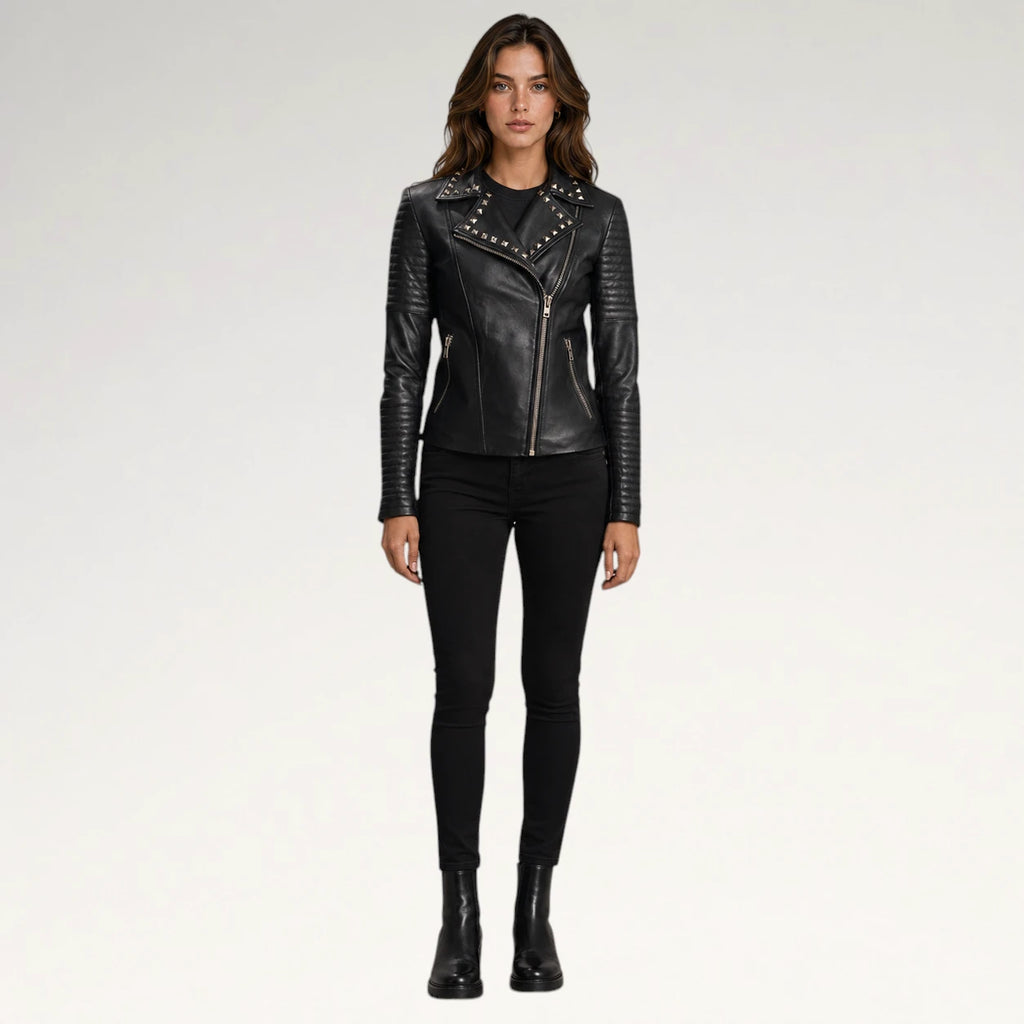 A woman modeling a black faux-leather moto jacket with silver studs and a diagonal zipper, paired with black skinny pants and black ankle boots, against a light gray studio background.