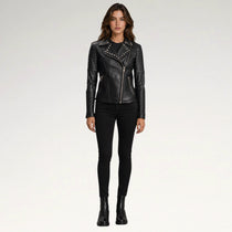 A woman modeling a black faux-leather moto jacket with silver studs and a diagonal zipper, paired with black skinny pants and black ankle boots, against a light gray studio background.
