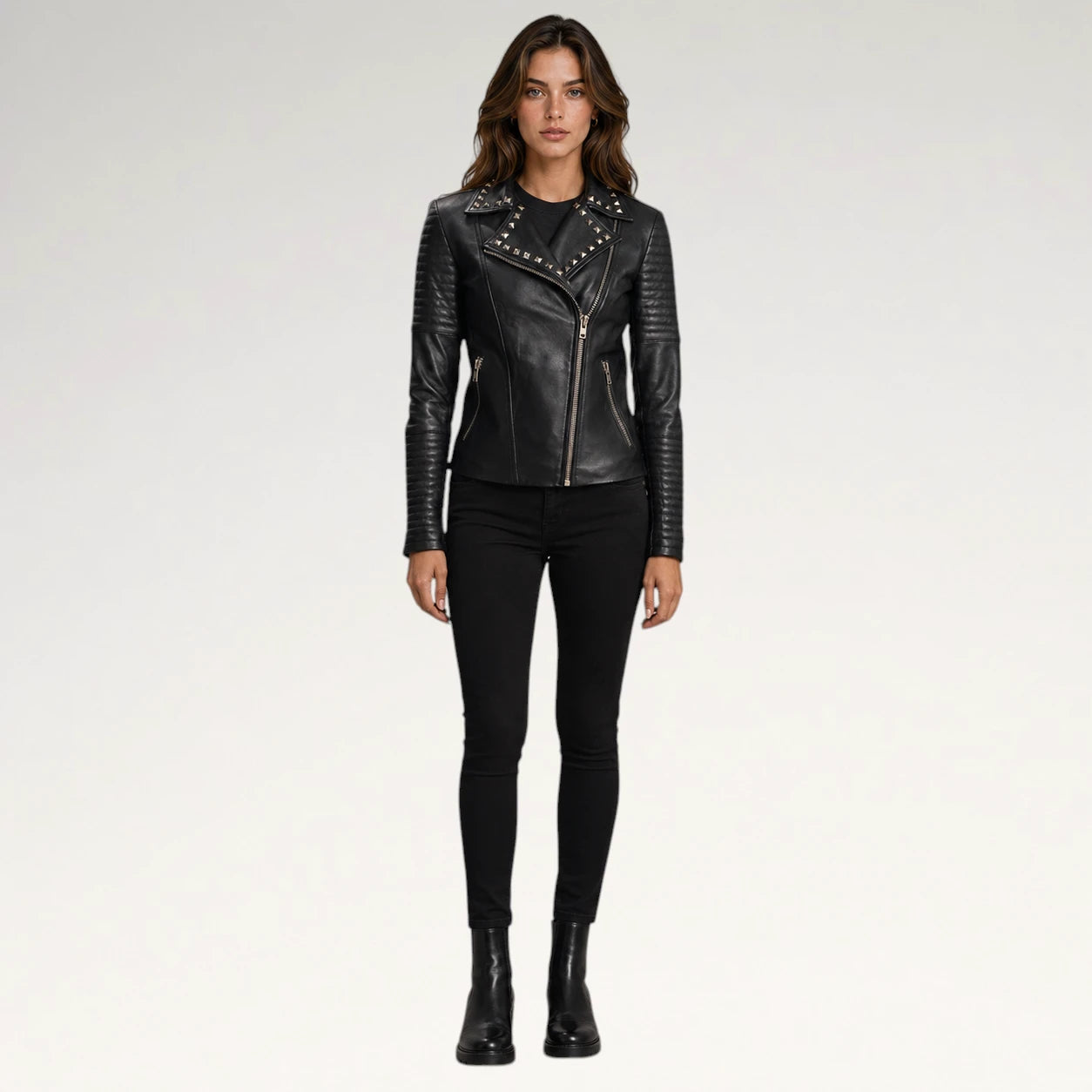 A woman modeling a black faux-leather moto jacket with silver studs and a diagonal zipper, paired with black skinny pants and black ankle boots, against a light gray studio background.