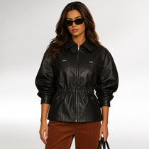 A woman modeling a black faux-leather jacket with a collar, front zipper, and gathered waist, paired with brown corduroy pants against a light gray studio background.
