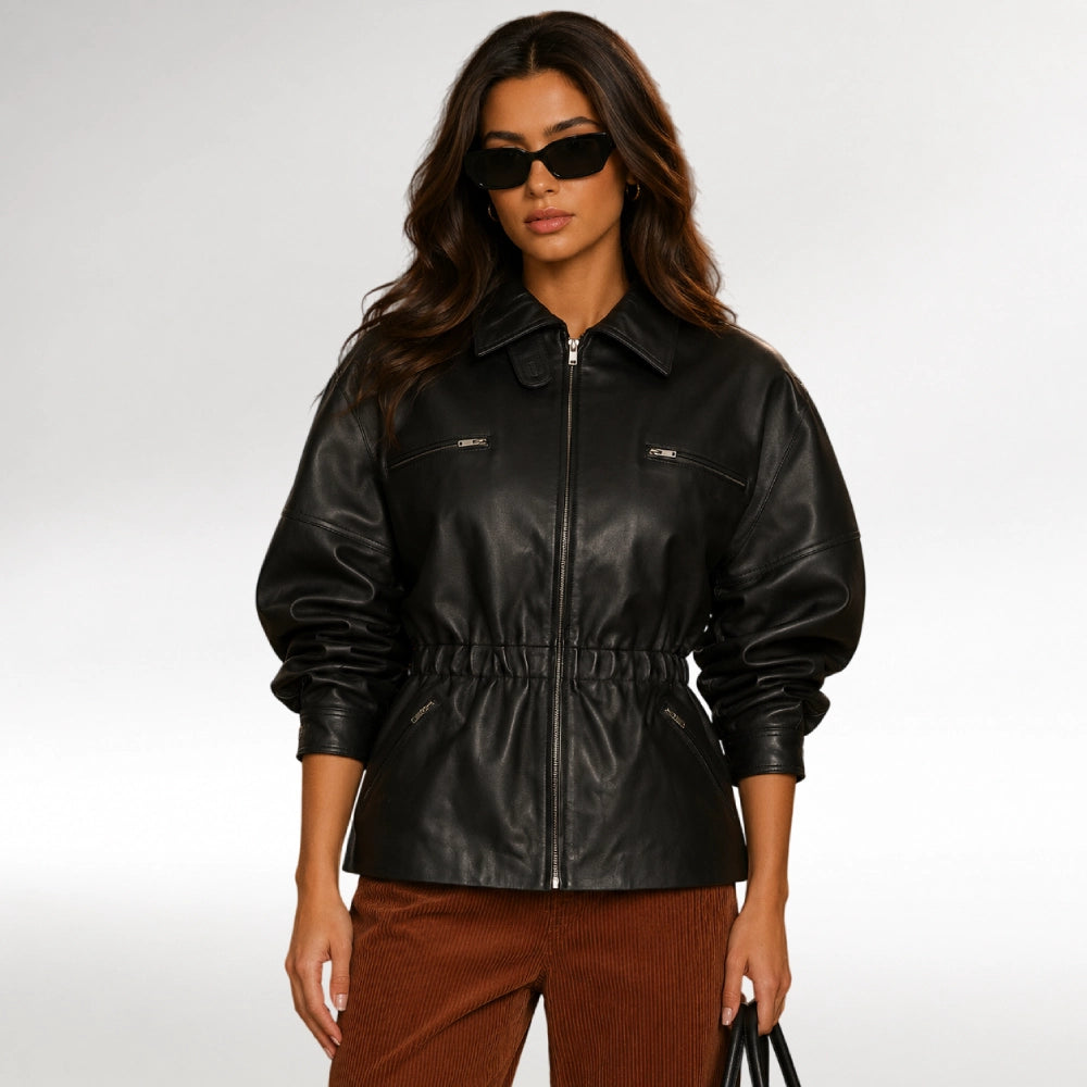 A woman modeling a black faux-leather jacket with a collar, front zipper, and gathered waist, paired with brown corduroy pants against a light gray studio background.